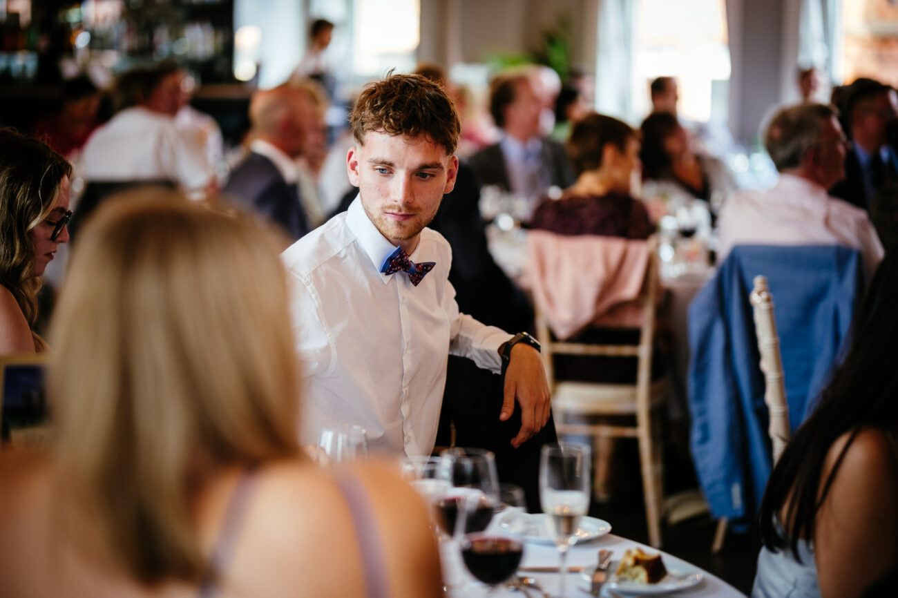 A man in a white shirt and bow tie sits at a table in a crowded dining room, looking to his right, with glasses of wine and plates of food visible on the tables.