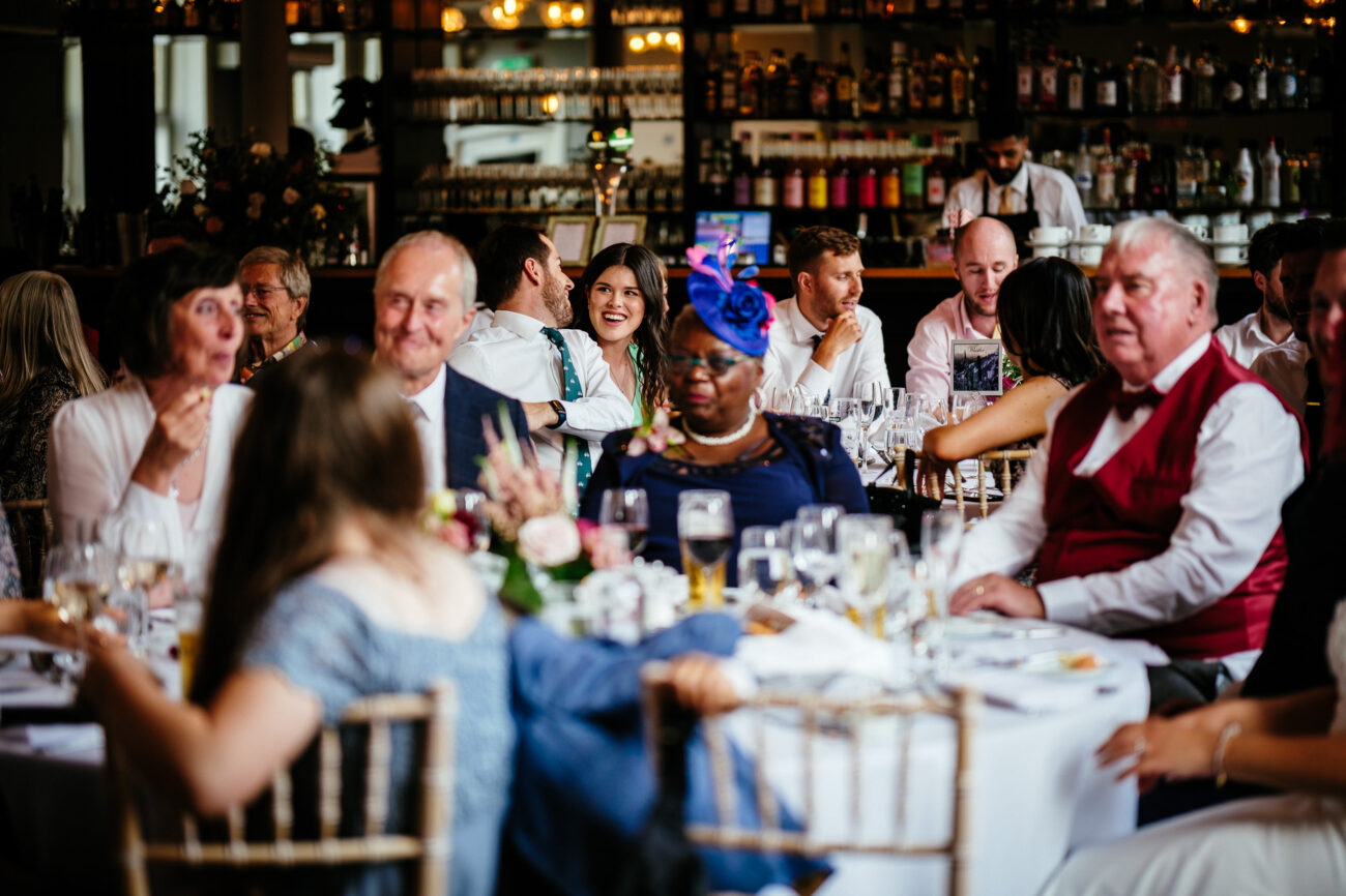 A group of people seated at round tables in a well-lit, elegant restaurant, engaged in conversation during a social gathering.