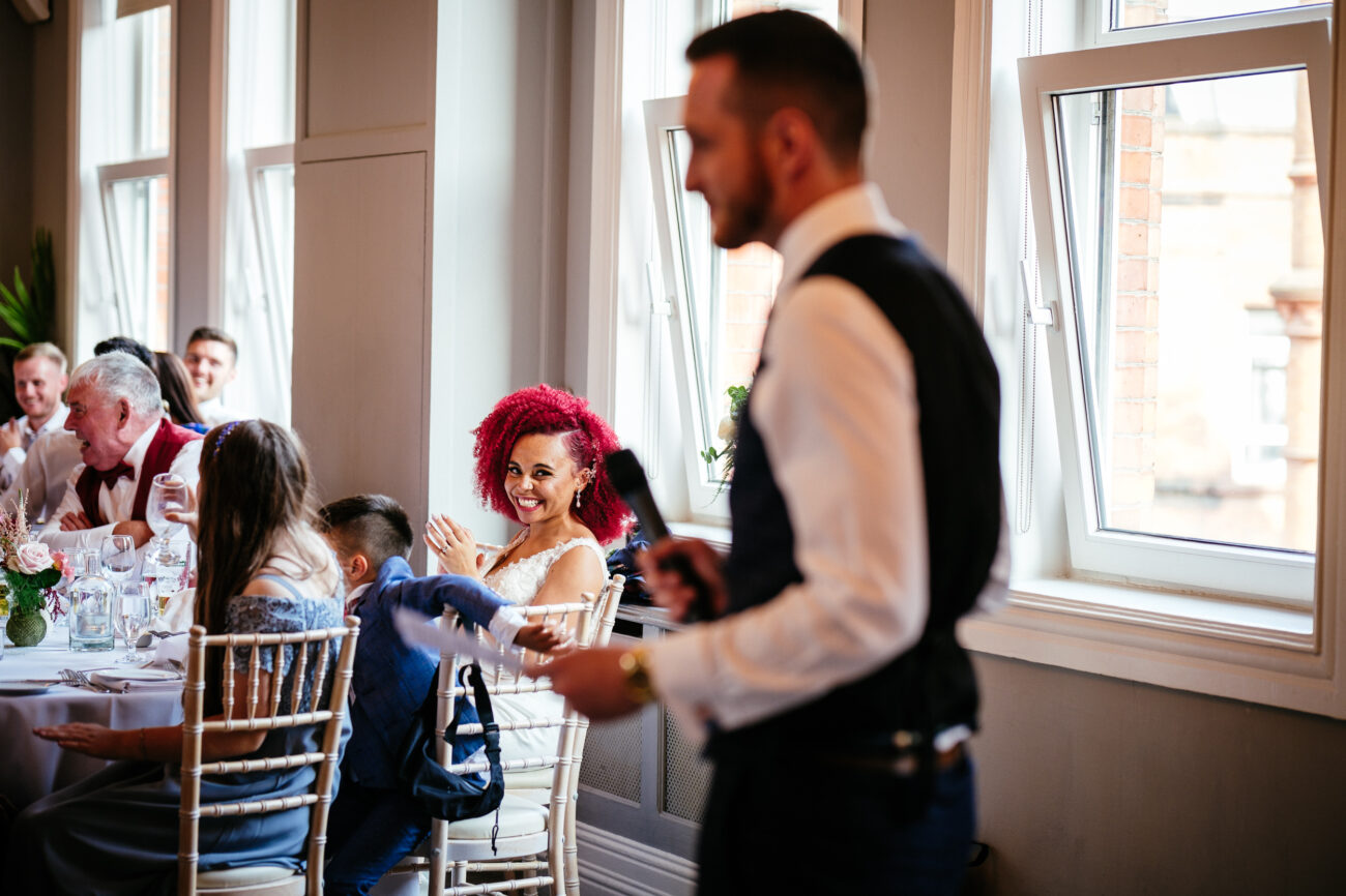 A man gives a speech in a well-lit room with large windows, while seated guests listen and smile.
