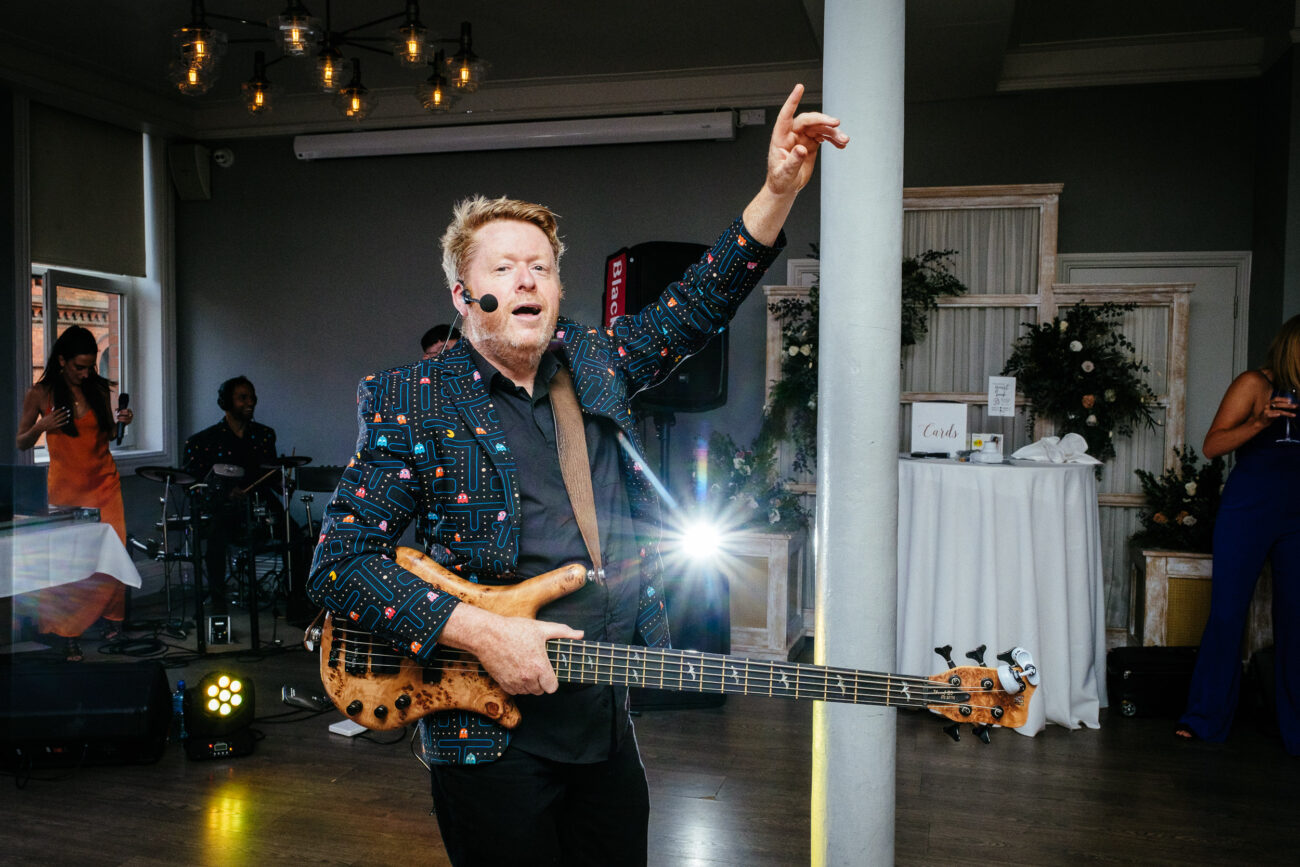 A musician wearing a patterned jacket plays a bass guitar and sings into a microphone, with another band member and various equipment visible in the background.