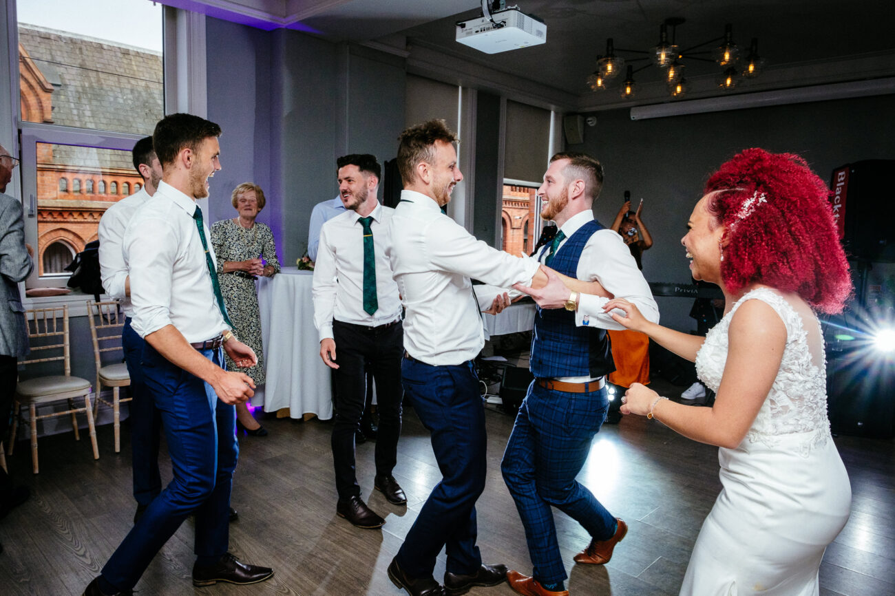 A group of people, mostly men in white shirts and ties, are dancing inside a room with wooden floors and large windows. A woman in a white dress with red curly hair joins them, smiling.