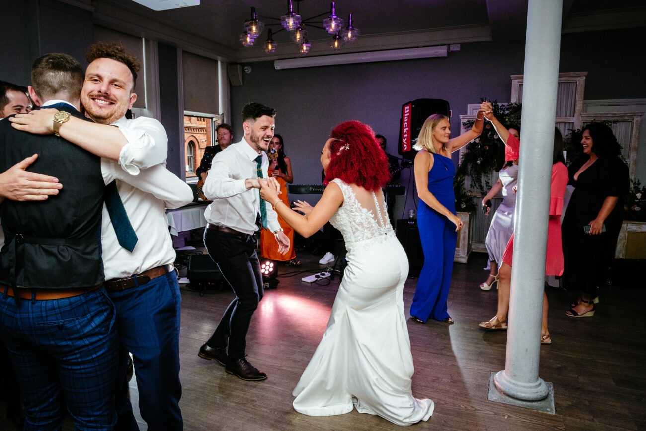 People are dancing and hugging in a well-lit room, wearing formal attire. A bride in a white dress is dancing with a man in a white shirt. Others are similarly engaged in joyful activity.