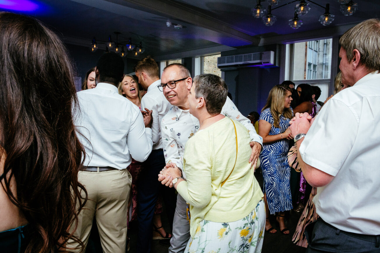A group of people dance and socialize in a well-lit room. A couple in the foreground, both smiling, dance together. Other guests are seen talking and dancing in the background.