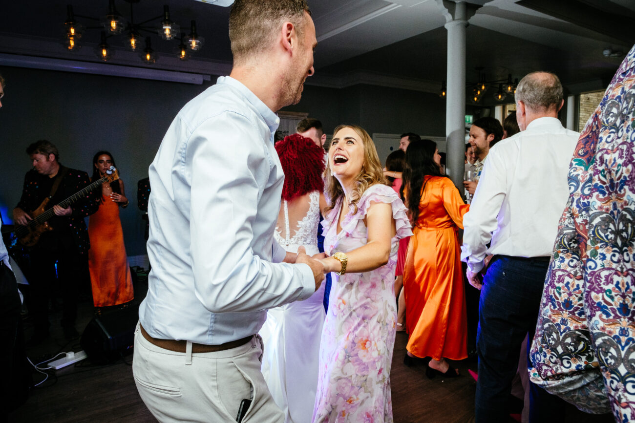 People are dancing and smiling at a joyous indoor event with a band playing in the background. The scene includes vibrant colors and warm lighting.