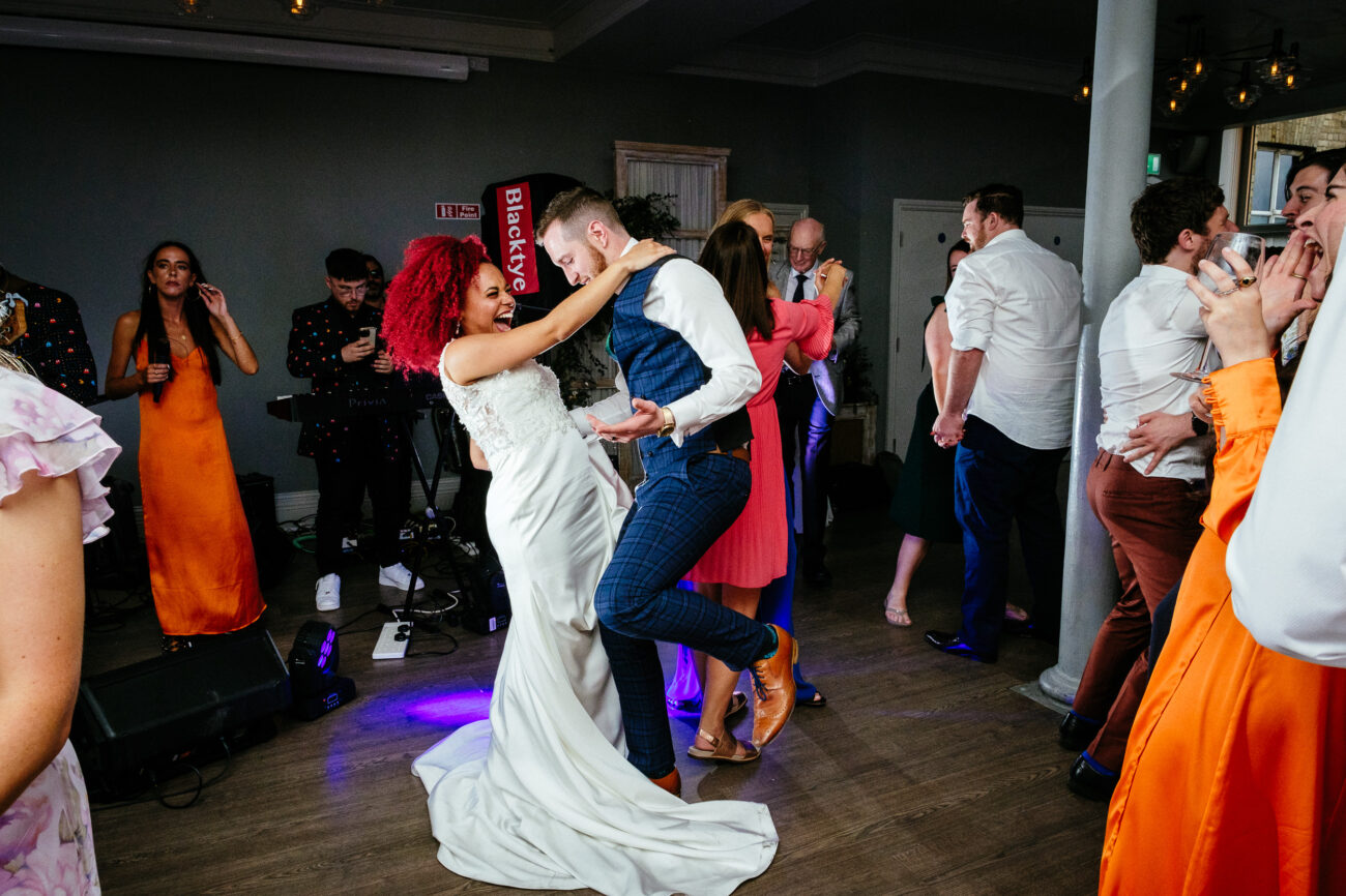 A couple, dressed in a white wedding gown and a blue suit, dance amidst a lively group of people inside a well-lit hall. Some individuals in the background cheer and applaud.