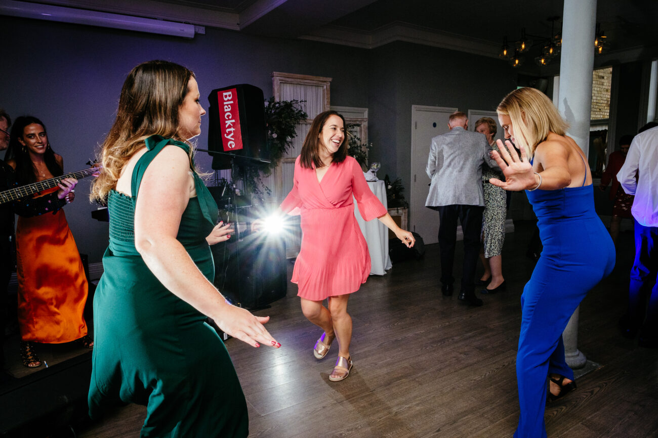 Three women dance energetically at a party in a room with dim lighting, while other attendees are visible in the background. One person in an orange dress appears to be singing or playing an instrument.