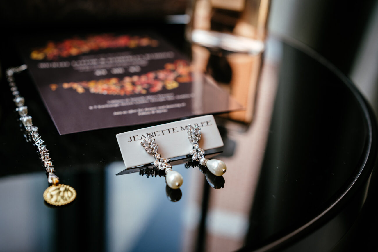 A pair of pearl and crystal earrings labeled "Jeanette Maree" is displayed on a black surface next to a bracelet and a floral wedding invitation.
