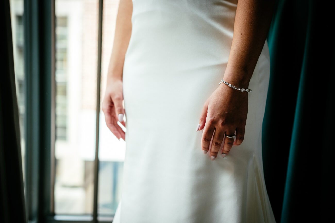 A person in a white dress stands near a window. Only their torso and hands are visible. They wear rings and a bracelet on their left hand.