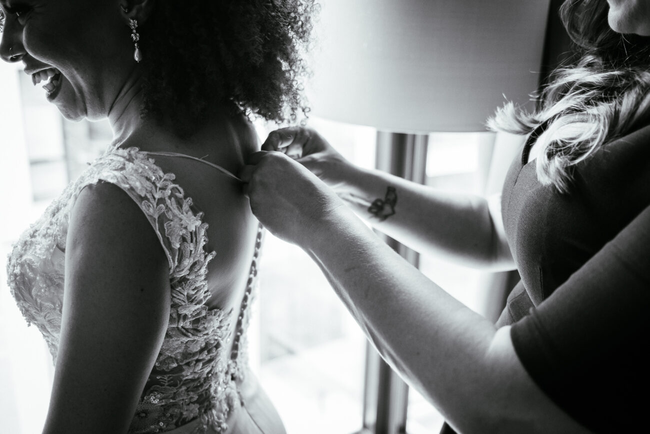 A woman assists a bride in fastening her dress near a window. The bride is wearing an intricate lace gown. The scene is captured in black and white.