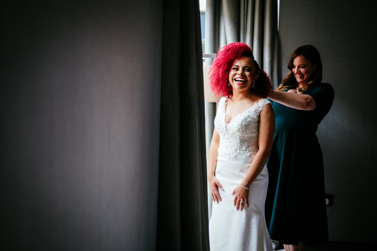 A bride in a white gown with bright pink hair smiles as a woman in a dark dress helps adjust her dress near a window.
