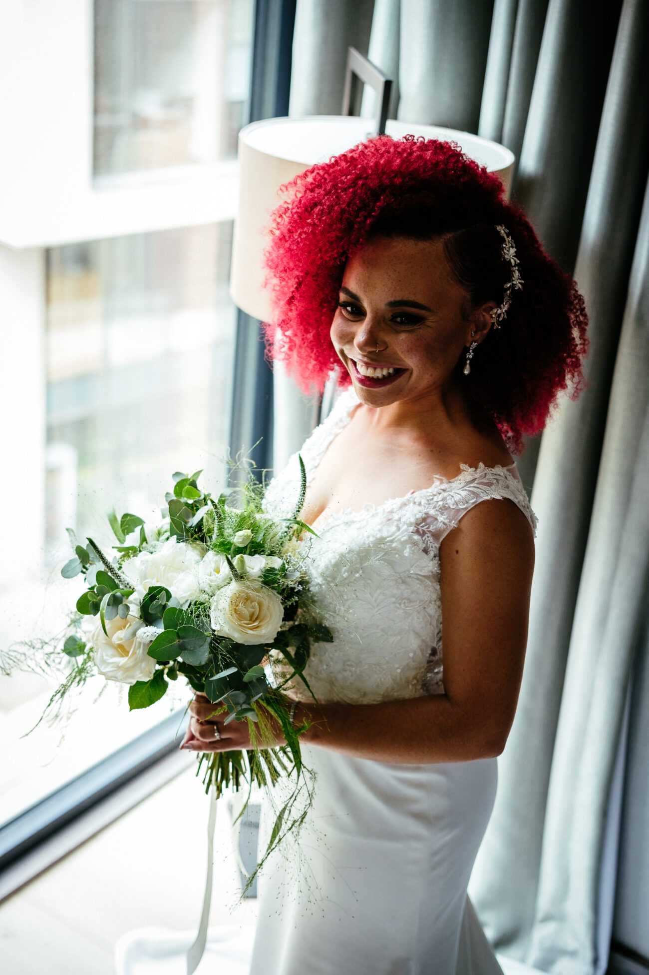 A bride with red curly hair and a sleeveless white gown holds a bouquet of white flowers while standing by a window with gray curtains.