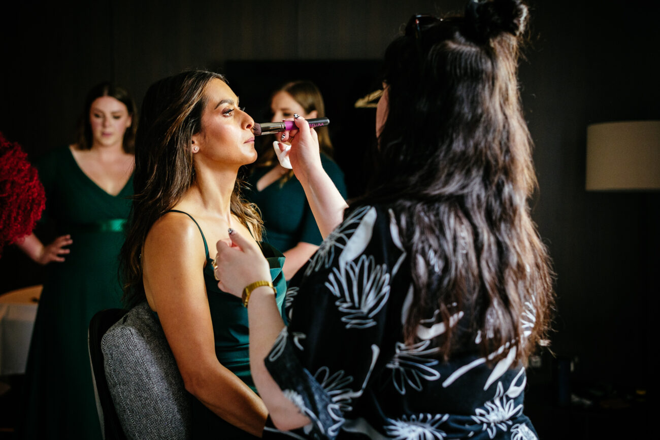 A woman sits as another woman applies makeup to her face in a dimly lit room. Three other women in green dresses stand in the background.
