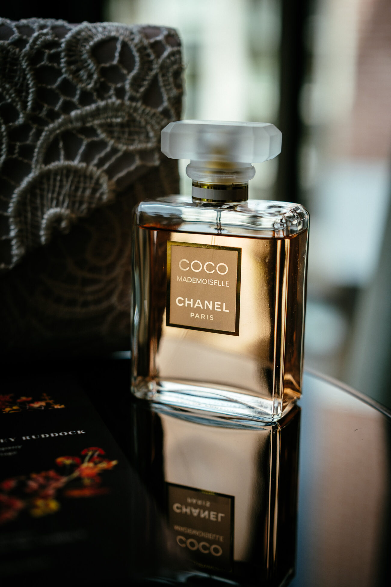 A bottle of Coco Mademoiselle Chanel perfume with a gold label sits on a reflective surface beside a lace fabric.