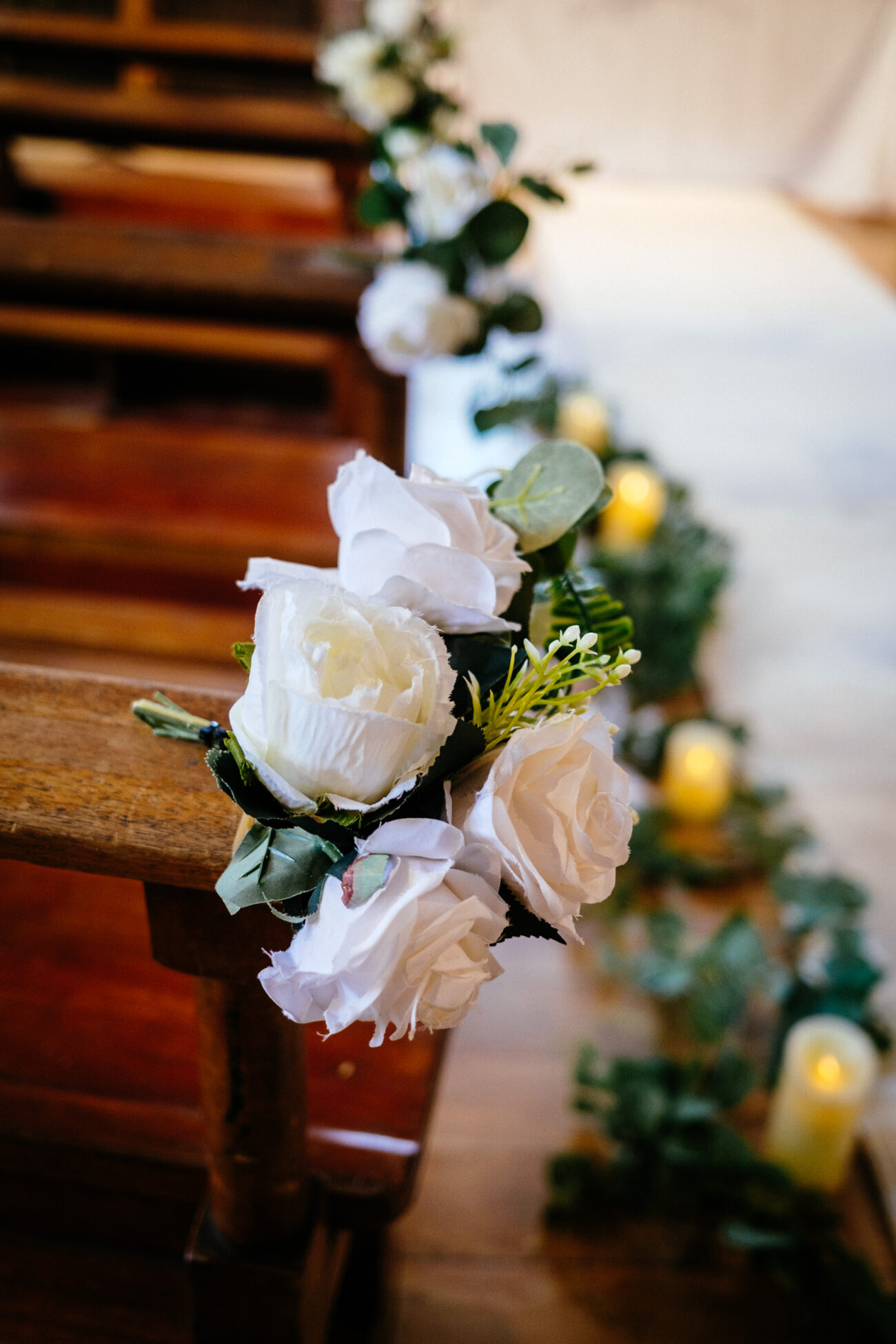 White floral decorations attached to wooden pews in an aisle setting with greenery and candles lining the aisle.