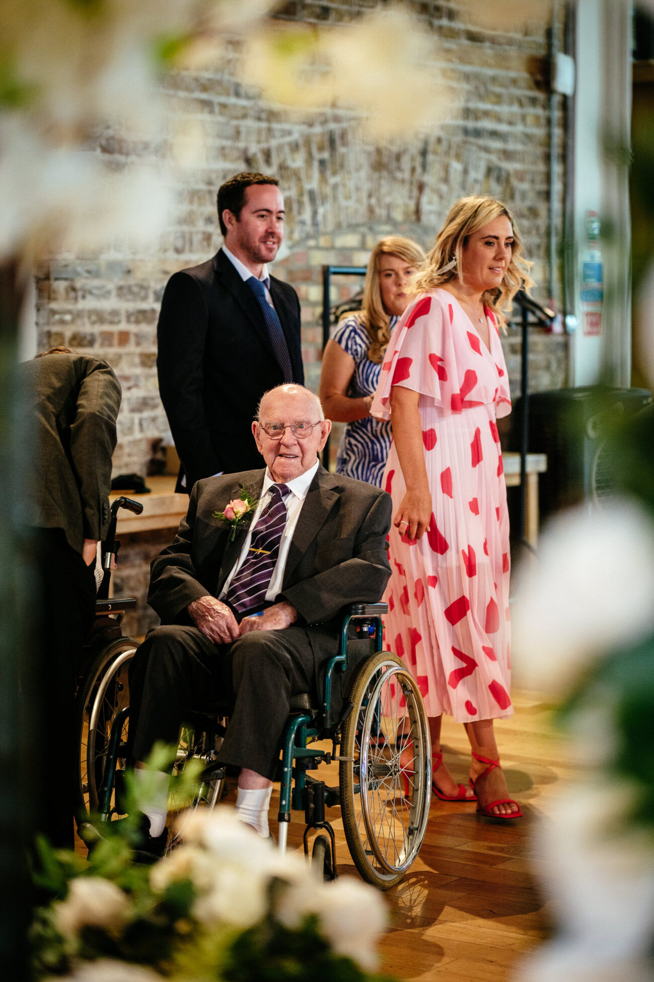An elderly man in a wheelchair wearing a suit and tie sits in the foreground. Behind him, three adults stand, including a woman in a pink dress with red hearts, against a brick wall.