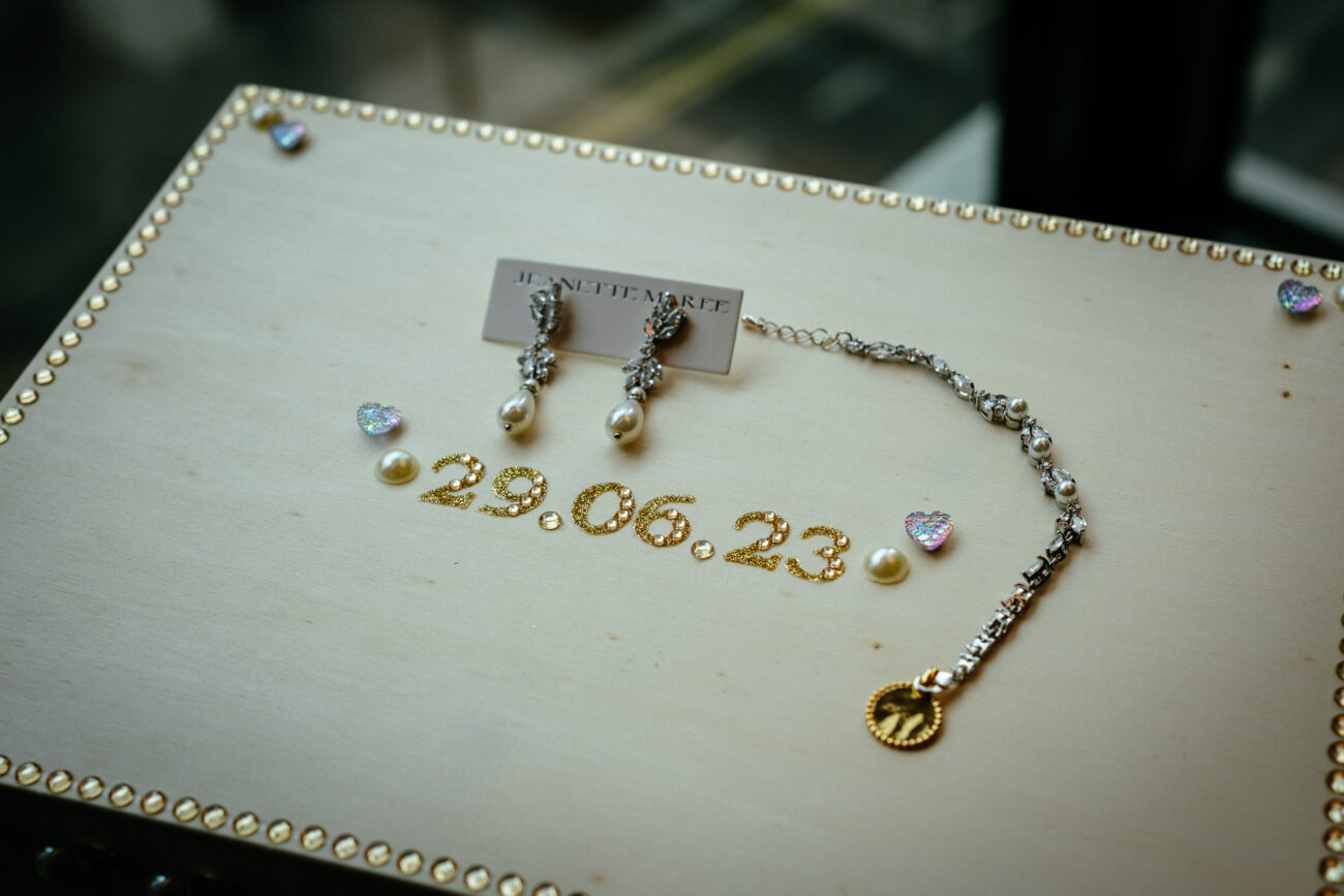 A wooden board displays a pair of pearl earrings and a beaded necklace with a gold pendant. The board is decorated with small hearts and the date "29.06.23.