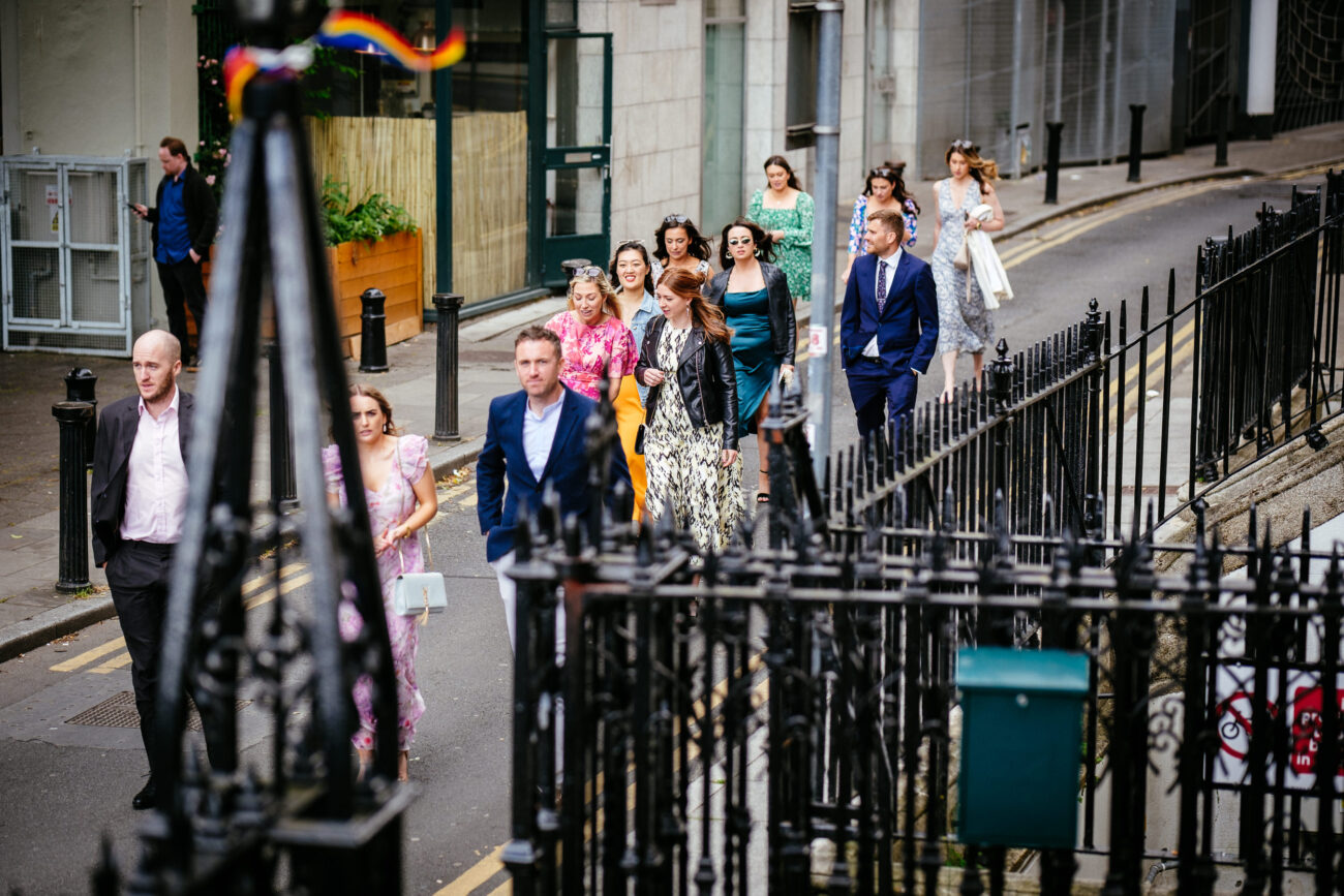 A formally dressed group of people, including men and women, walk along a city street near a wrought iron fence. Some women wear hats.
