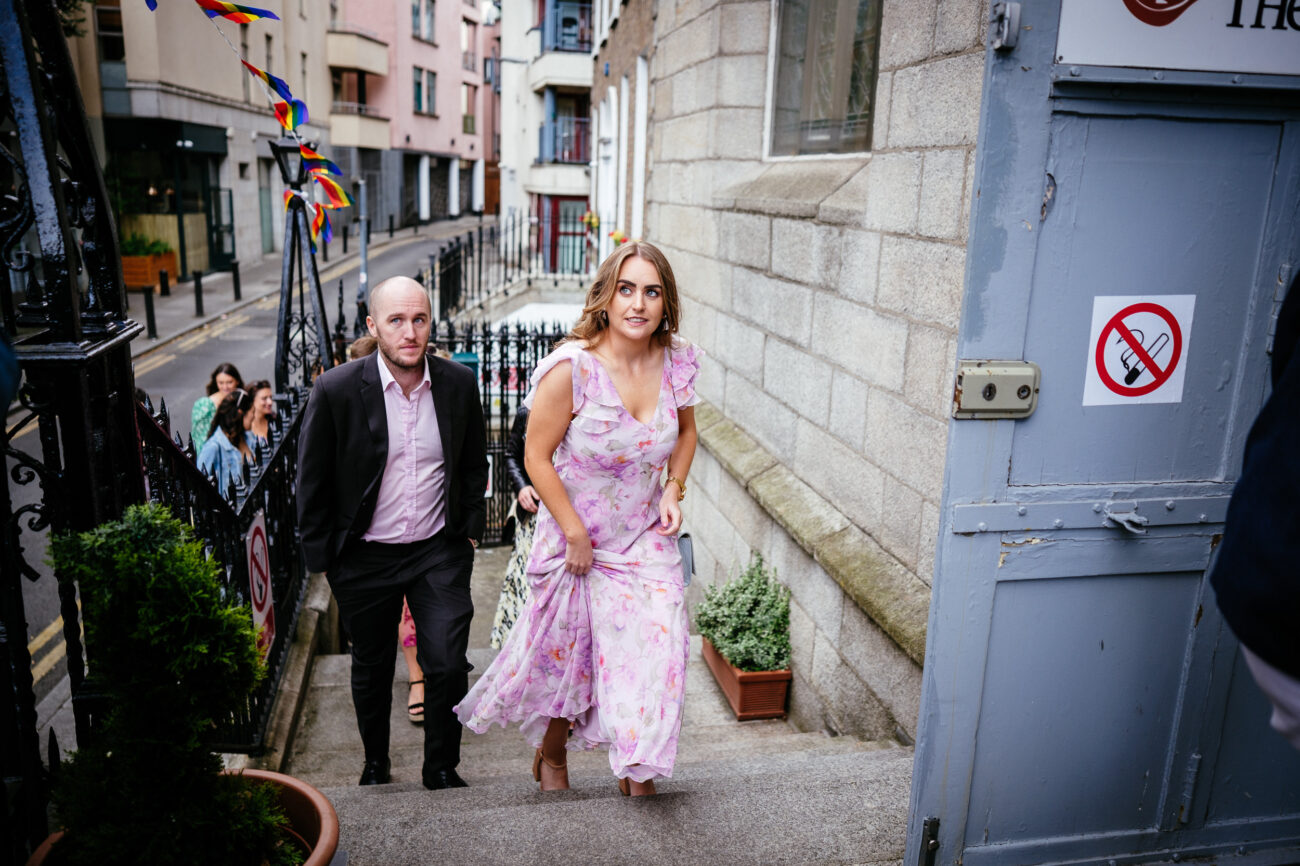A woman in a floral dress and a man in a suit ascend stone stairs beside a blue door with a no-smoking sign, with people and colorful flags visible in the background.