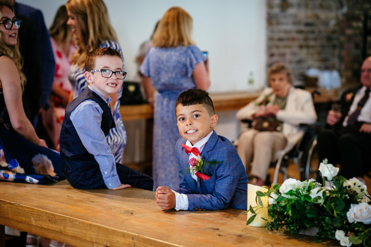 Two boys in suits sitting on a wooden table, surrounded by people at an indoor event. Floral arrangements are visible on the table.
