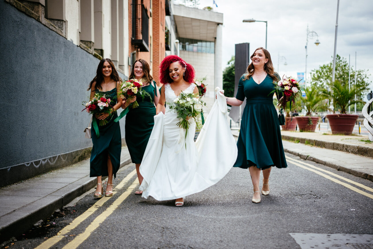 A bride in a white dress walks down a street with three bridesmaids in dark green dresses, all holding bouquets.