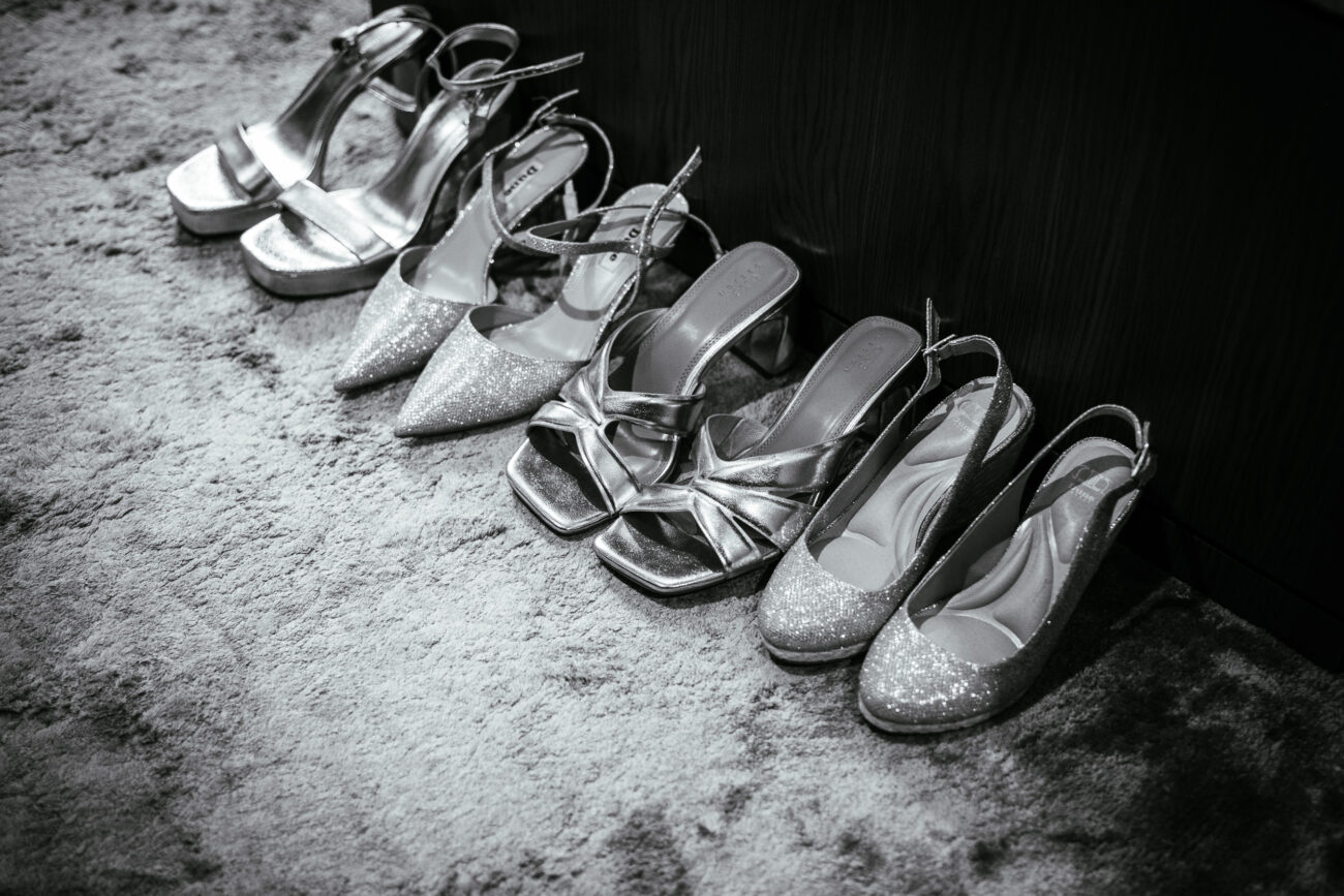 A row of women's high-heeled shoes in various styles, including strappy, pointed, and platform, arranged on a carpeted floor. The image is in black and white.