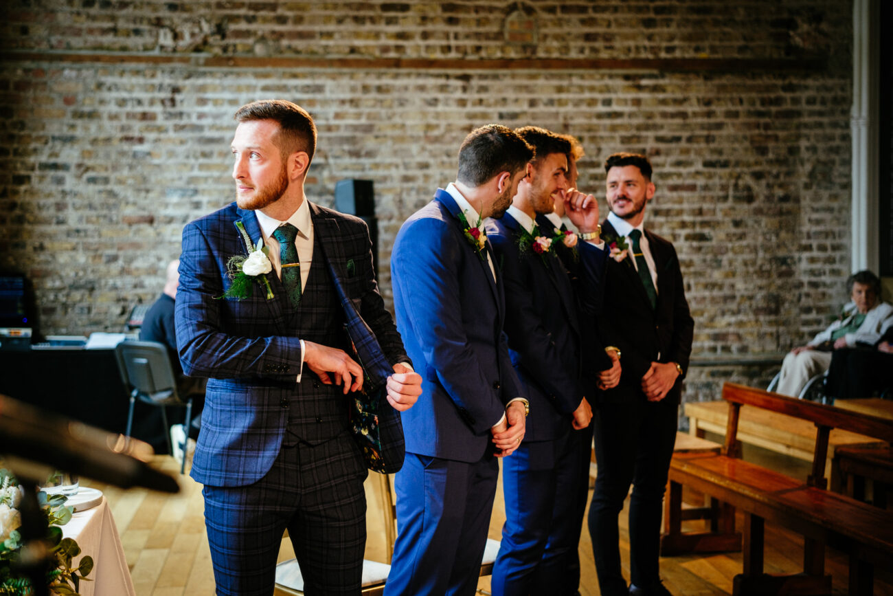 Four men in suits stand in a row in front of a brick wall, with one looking to the side and the others interacting with each other. A seated person and wooden benches are visible in the background.