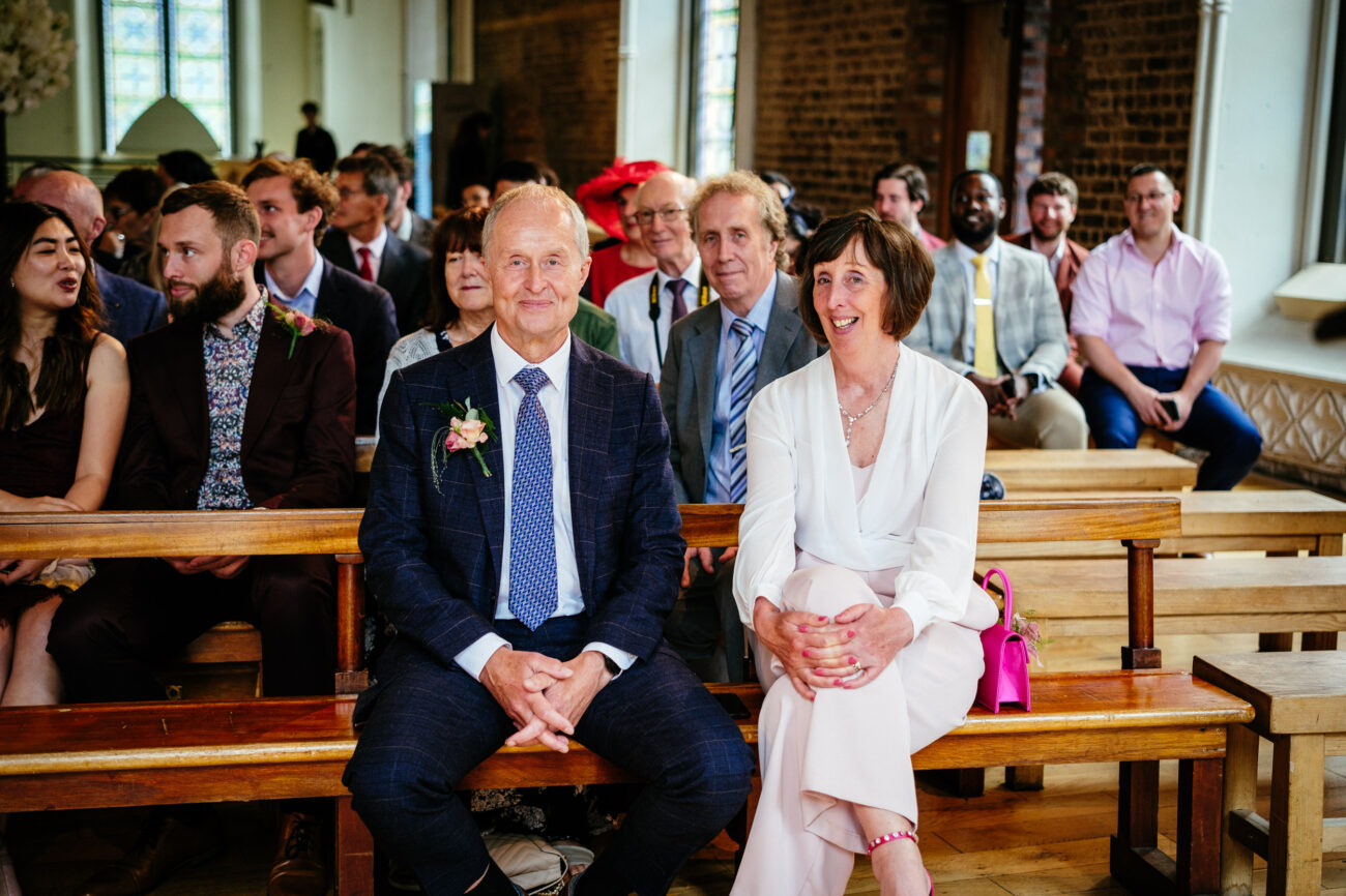 A group of people, dressed in formal attire, are seated on wooden benches inside a building with large windows and brick walls. A man and a woman sit at the front, smiling.