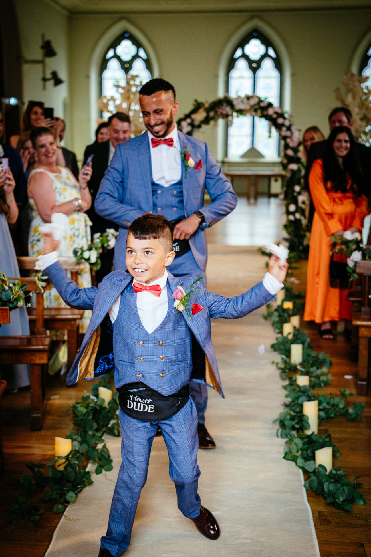 A young boy in a blue suit with a fanny pack that says "Ring Security" excitedly walks down the aisle of a decorated venue, followed by a smiling man in a matching blue suit. Guests are seated and watching.