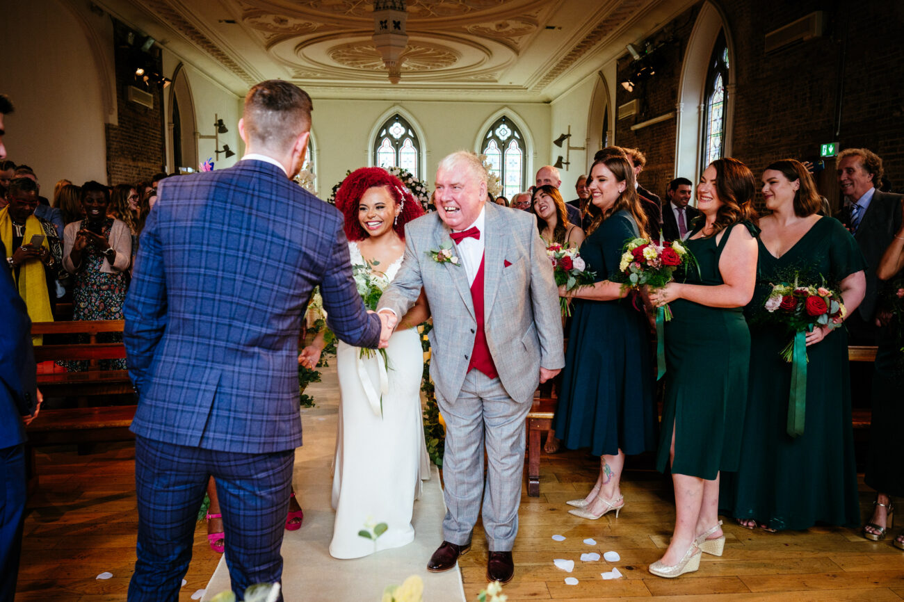 A bride and groom shake hands with an elder man in a gray suit and red bowtie inside a decorated church, surrounded by bridesmaids in green dresses and guests.