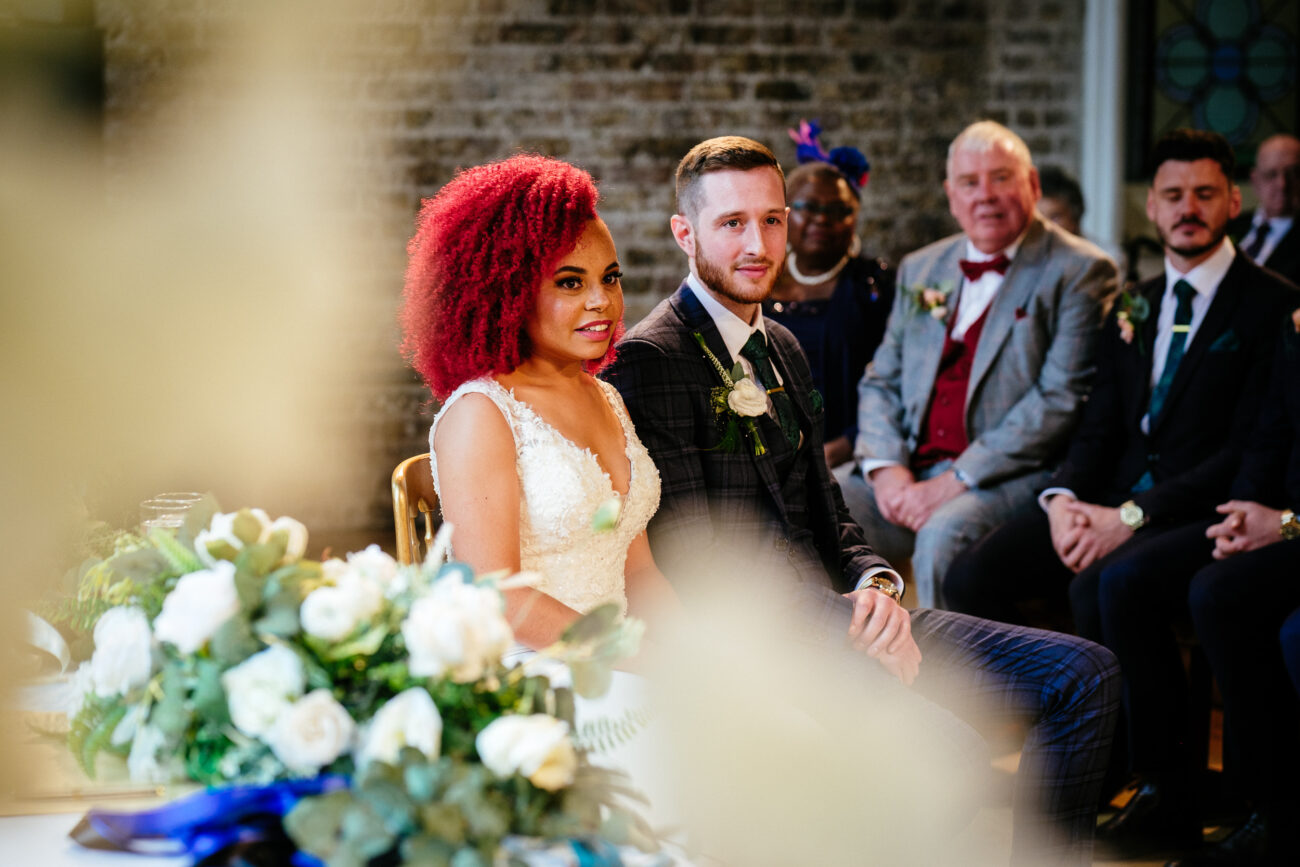 A couple sits during a wedding ceremony. The bride wears a white dress and has bright red hair. The groom is in a dark suit with a boutonniere. Guests are seated behind them. Floral decor is in the foreground.