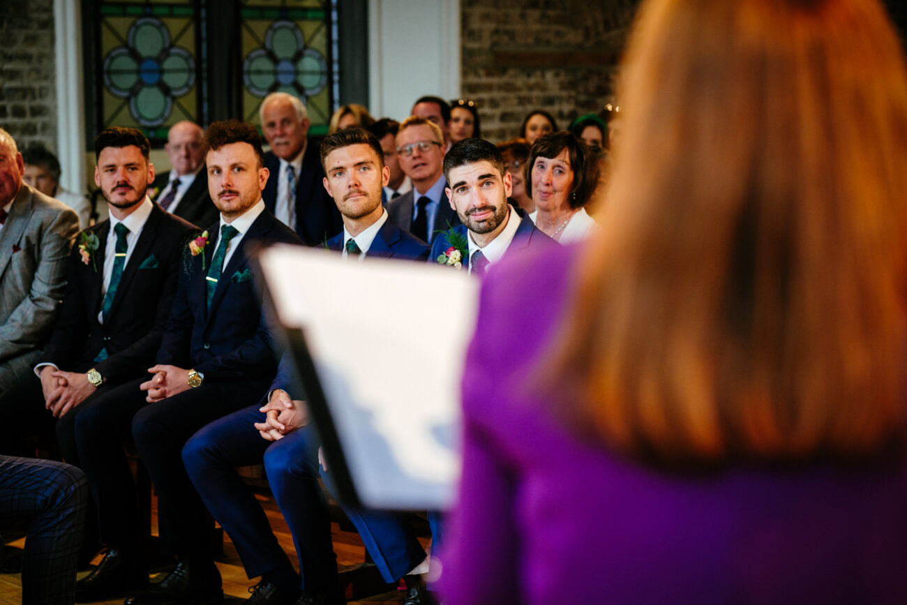 A group of people seated indoors, dressed formally. Men in suits are seated in the front row, listening attentively to a speaker, who is in focus from behind. Stained glass windows are visible in the background.