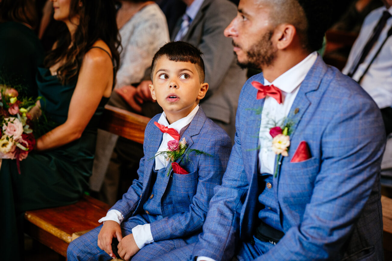 A boy and a man, both in blue suits with red bow ties, sit on a wooden bench at an indoor event. The boy looks up while the man looks forward. Other attendees are seen in the background.