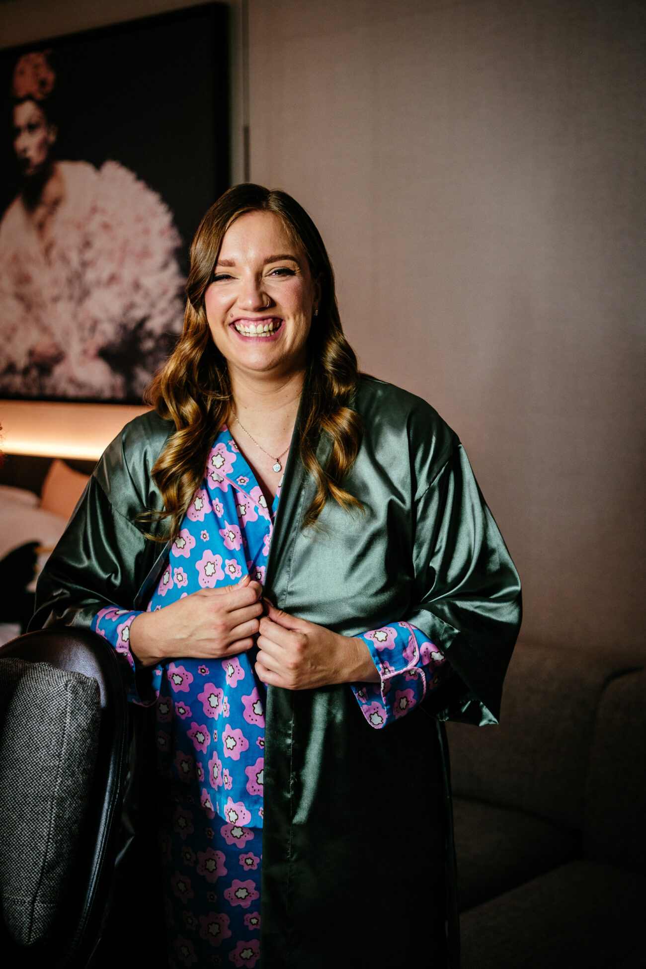 A woman with long, wavy hair wearing a green satin robe and a pink floral gown smiles while standing indoors in front of a framed photograph.