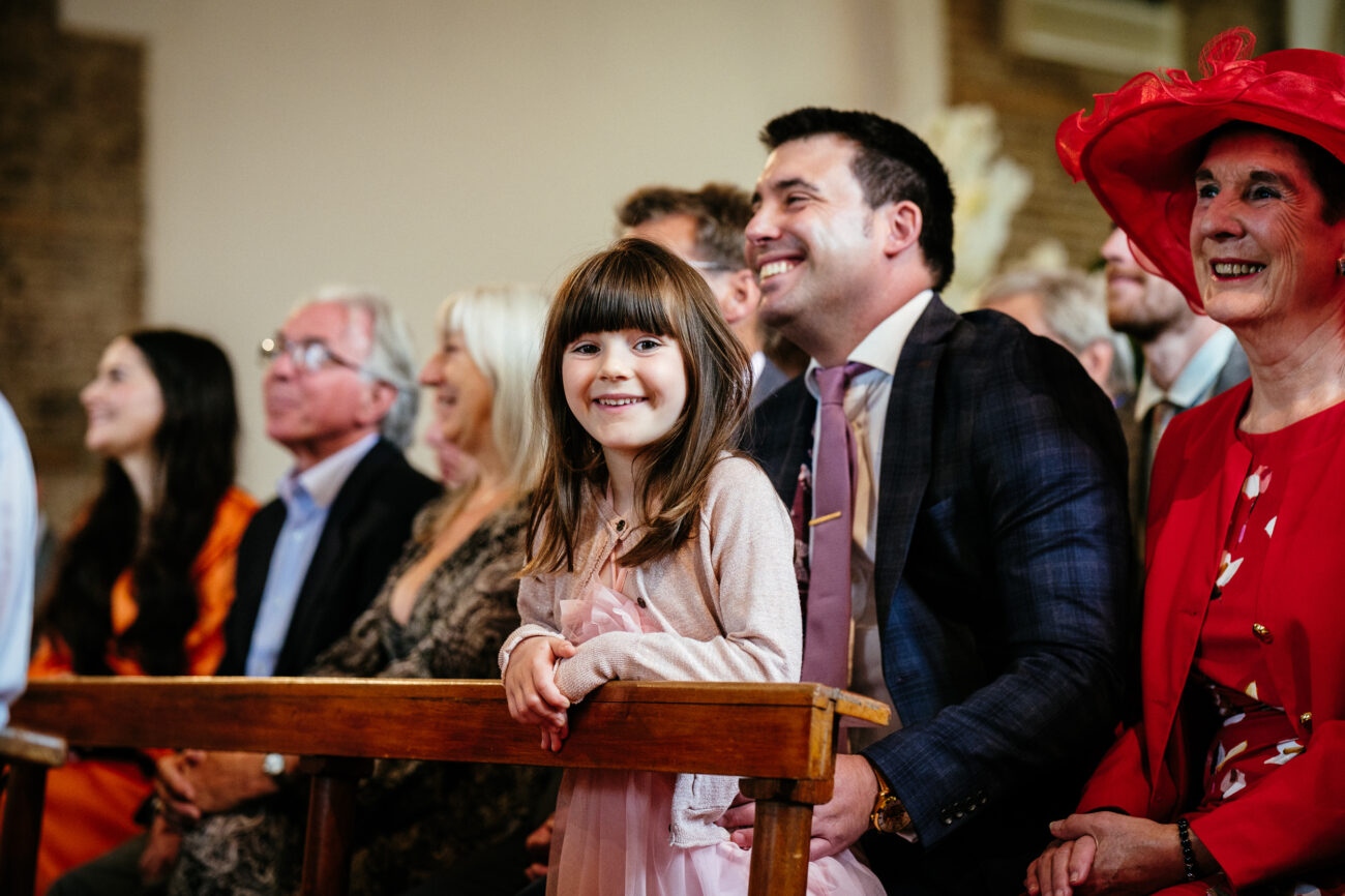A group of people, including a child in a pink dress, are seated inside a church. A person in a bright red hat sits at the right edge. The atmosphere appears joyful.
