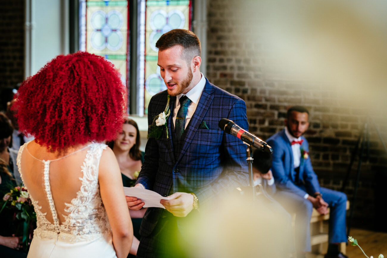A man in a blue plaid suit reads from a paper while a woman in a white dress stands facing him in a room with a stained glass window and brick wall. Seated guests are visible in the background.