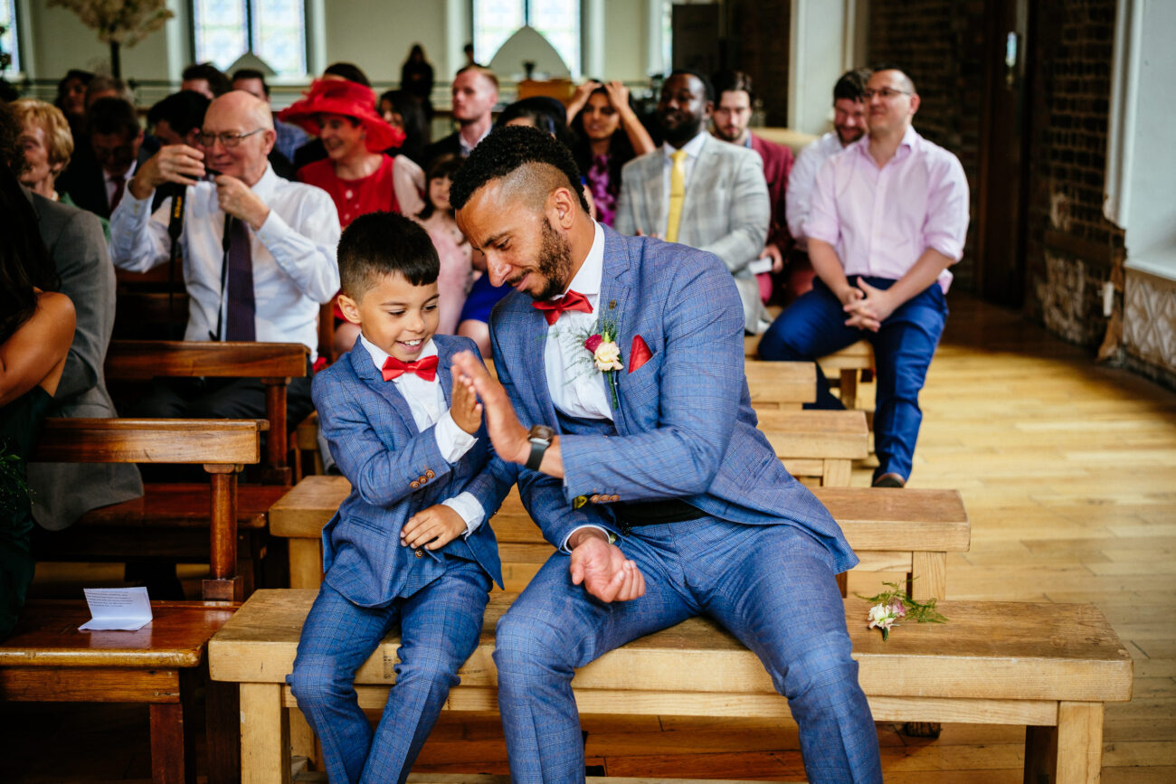 A man and a boy, both in matching blue suits with red bow ties, share a high-five while seated on a wooden bench, surrounded by other seated people in an indoor setting.