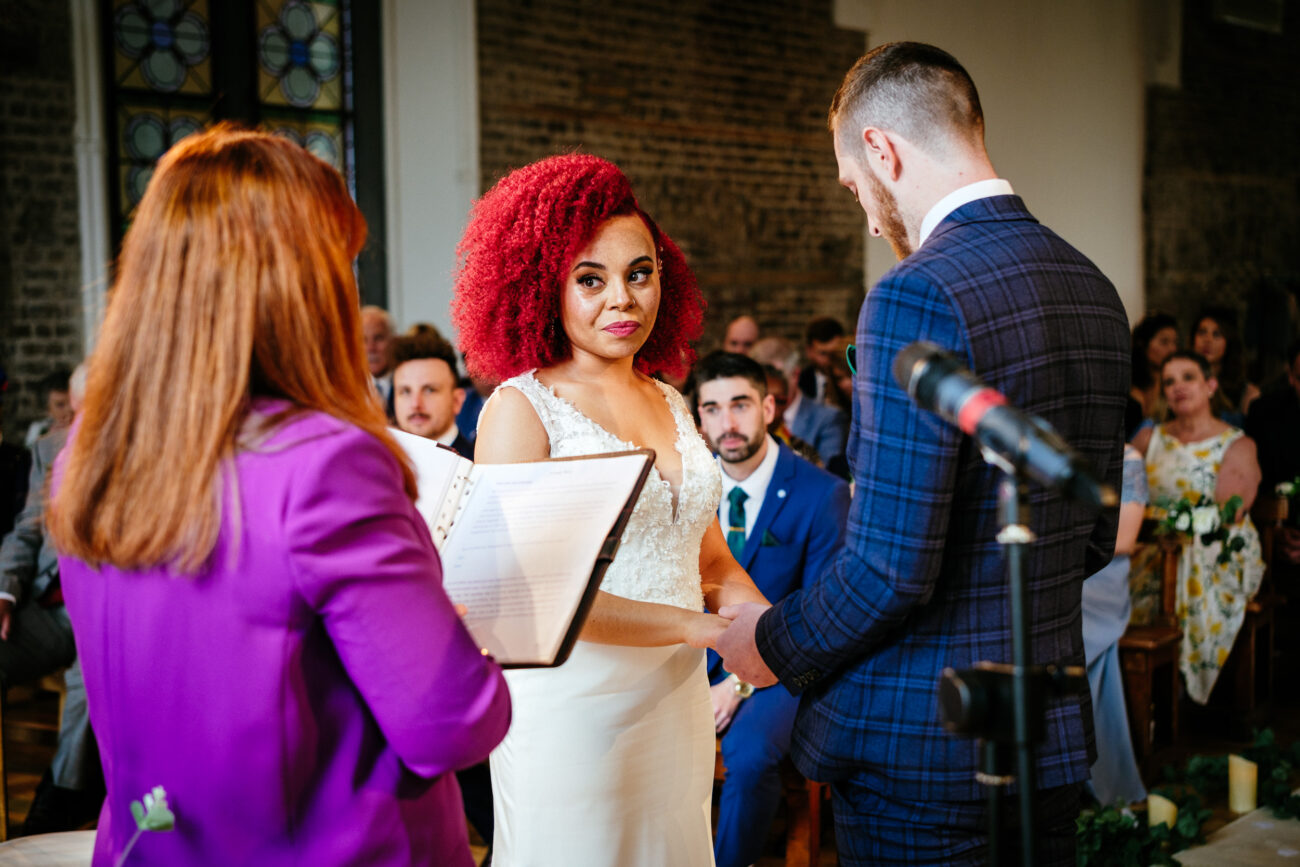 A couple holds hands and exchanges vows during a wedding ceremony officiated by a woman with red hair. Guests are seated in the background.
