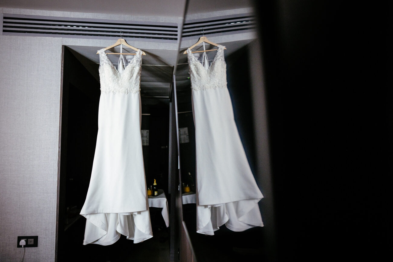 A white wedding dress with lace details hangs on a hanger, reflected in a mirror.