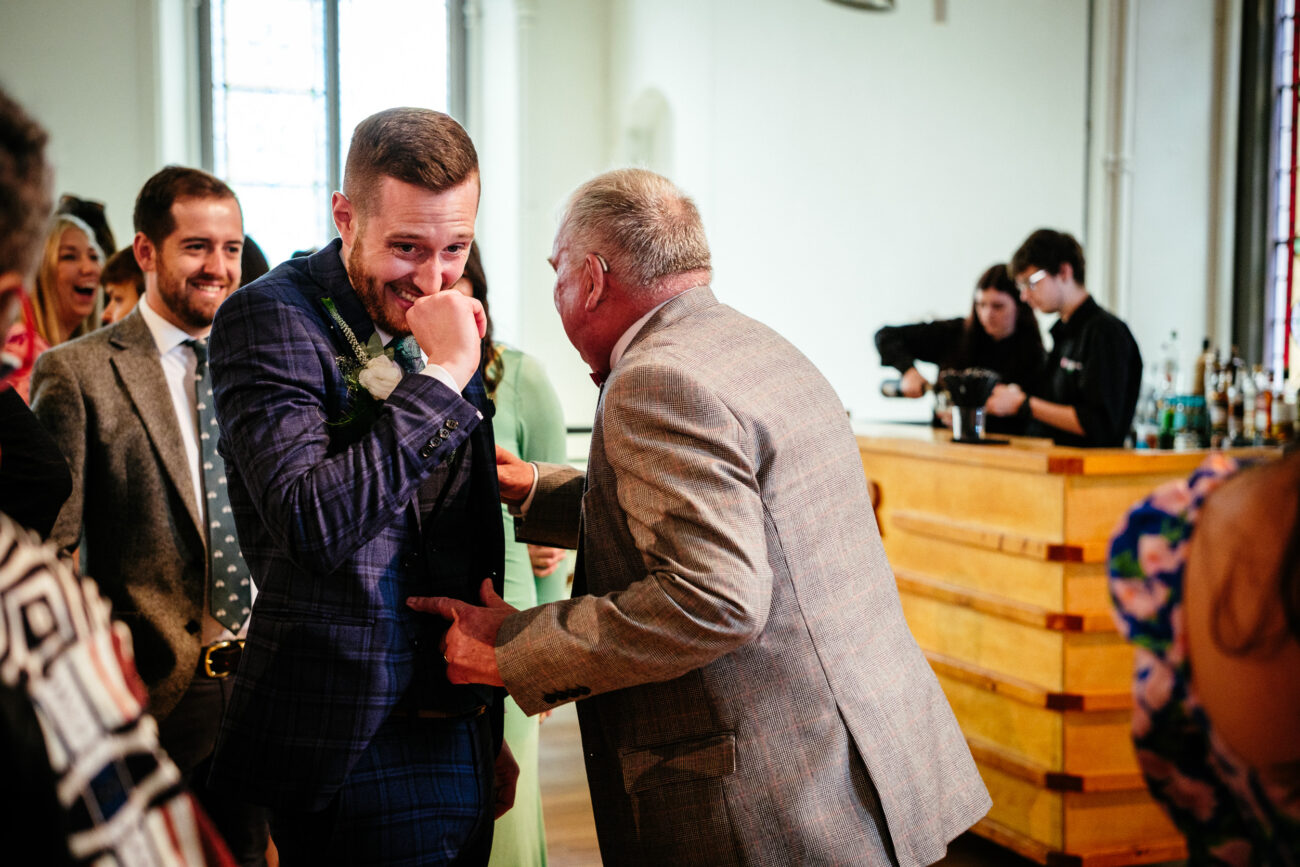 Two men are laughing and interacting at an indoor event. One is dressed in a blue checked suit, and the other in a gray suit. Other people and a bar are visible in the background.