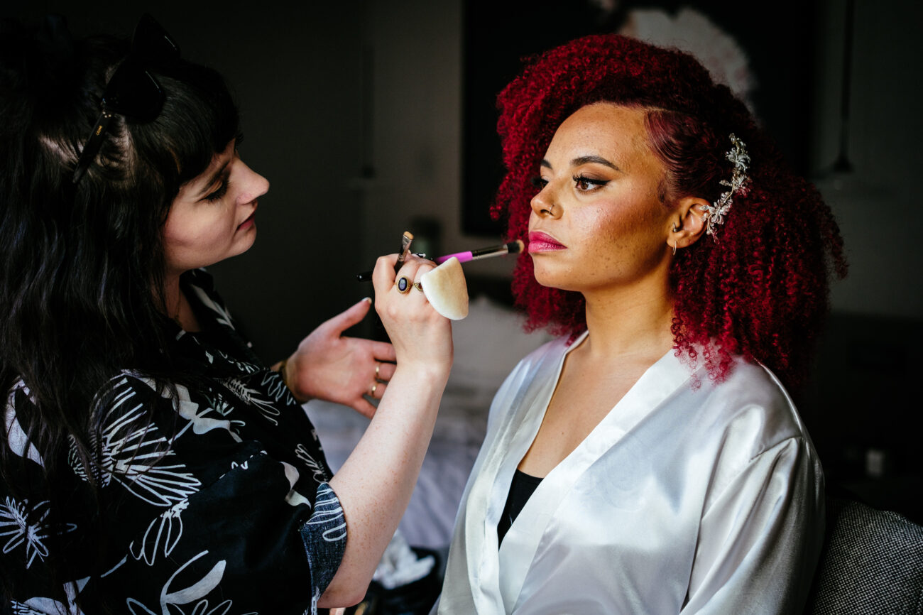 A woman with curly red hair, wearing a white satin robe, gets her makeup done by a makeup artist using a brush.