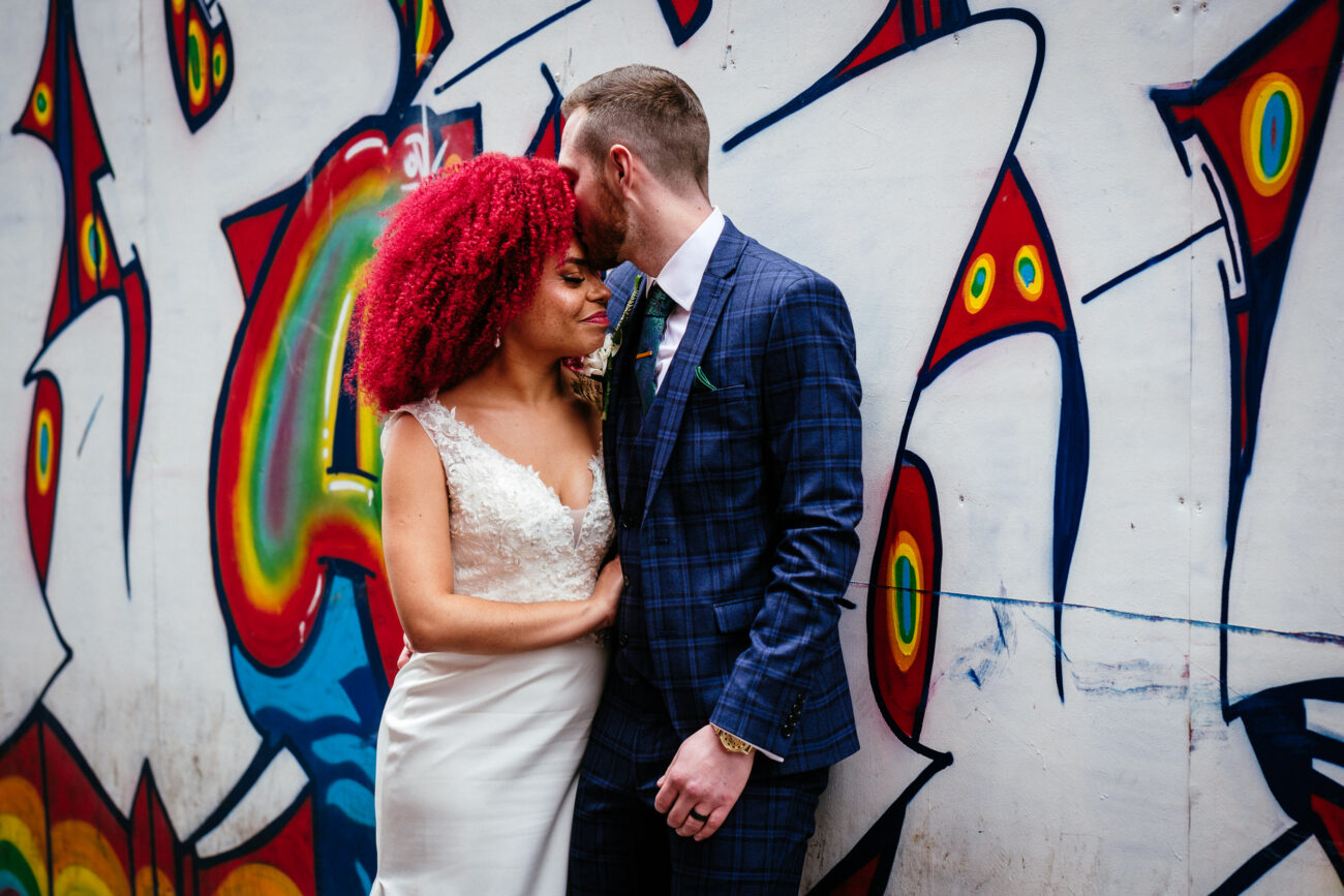 A couple stands close together with the man kissing the woman's forehead. They are in front of a colorful graffiti wall. The woman wears a white dress, and the man is in a blue checkered suit.