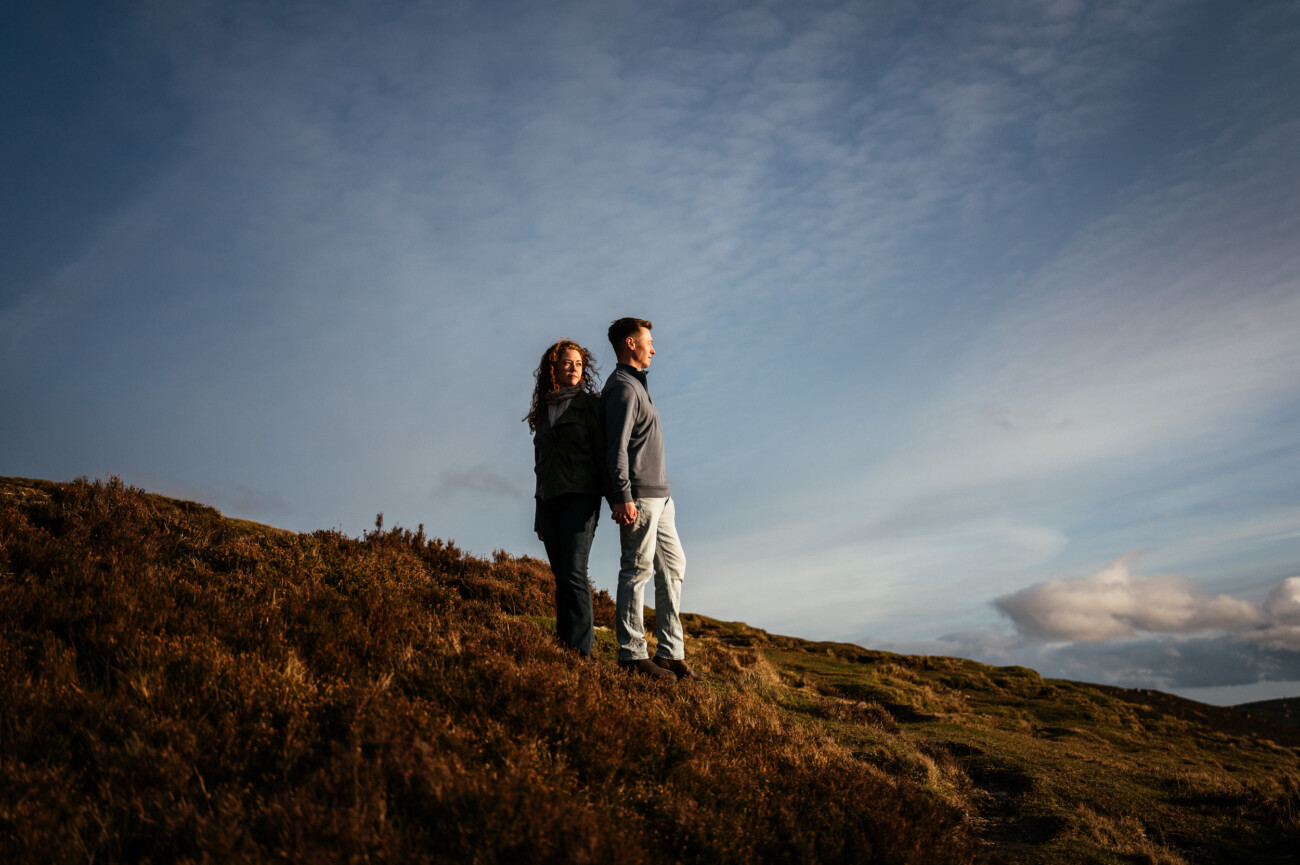 Two people stand side by side on a grassy hill under a blue sky with scattered clouds, looking into the distance—capturing a perfect moment at a top engagement shoot location in Dublin.