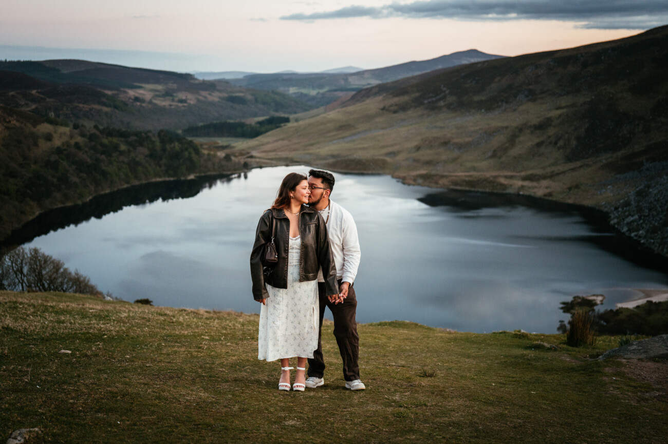 A couple stands close together on a grassy hillside overlooking a large, calm lake surrounded by rolling hills at sunset—truly a top engagement shoot location in Dublin.