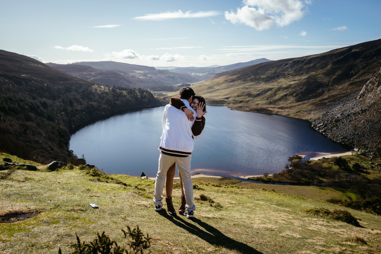 Two people embrace on a grassy hill overlooking a lake surrounded by mountains under a partly cloudy sky—a top engagement shoot location in Dublin.