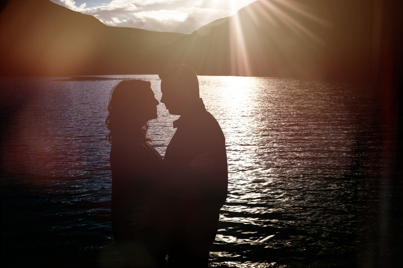Silhouette of a couple standing close together by a lake during sunset, with mountains in the background and sunlight streaming through the clouds.