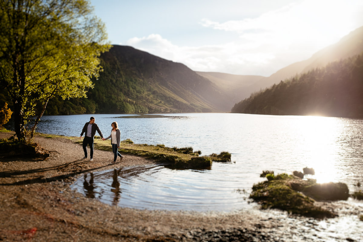 A person and a child walk hand in hand along a lakeshore path surrounded by mountains and trees under a bright sun for their engagement photos in Ireland.
