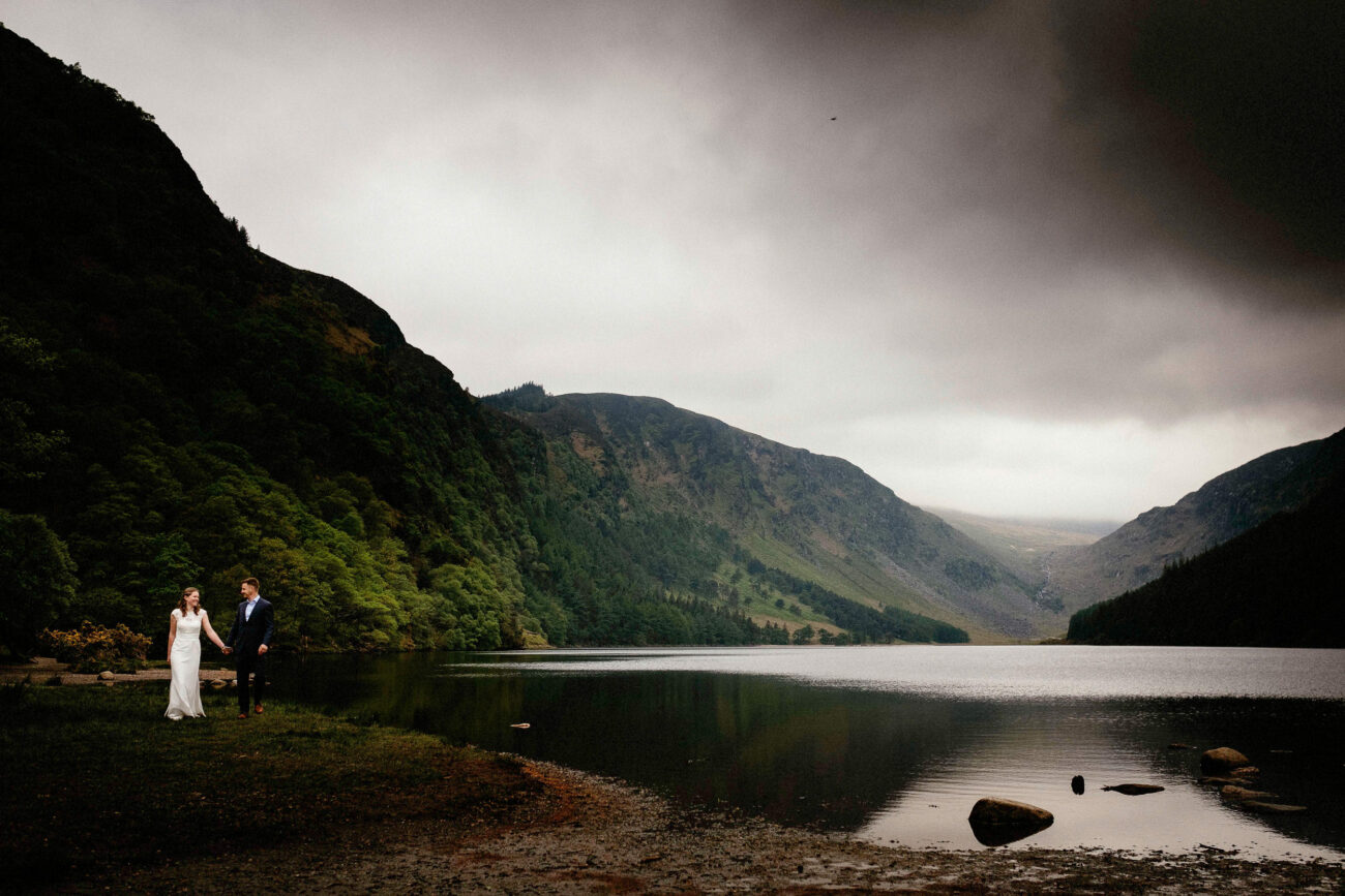 A couple in wedding attire stand on a grassy shore beside a lake, surrounded by mountains under an overcast sky.