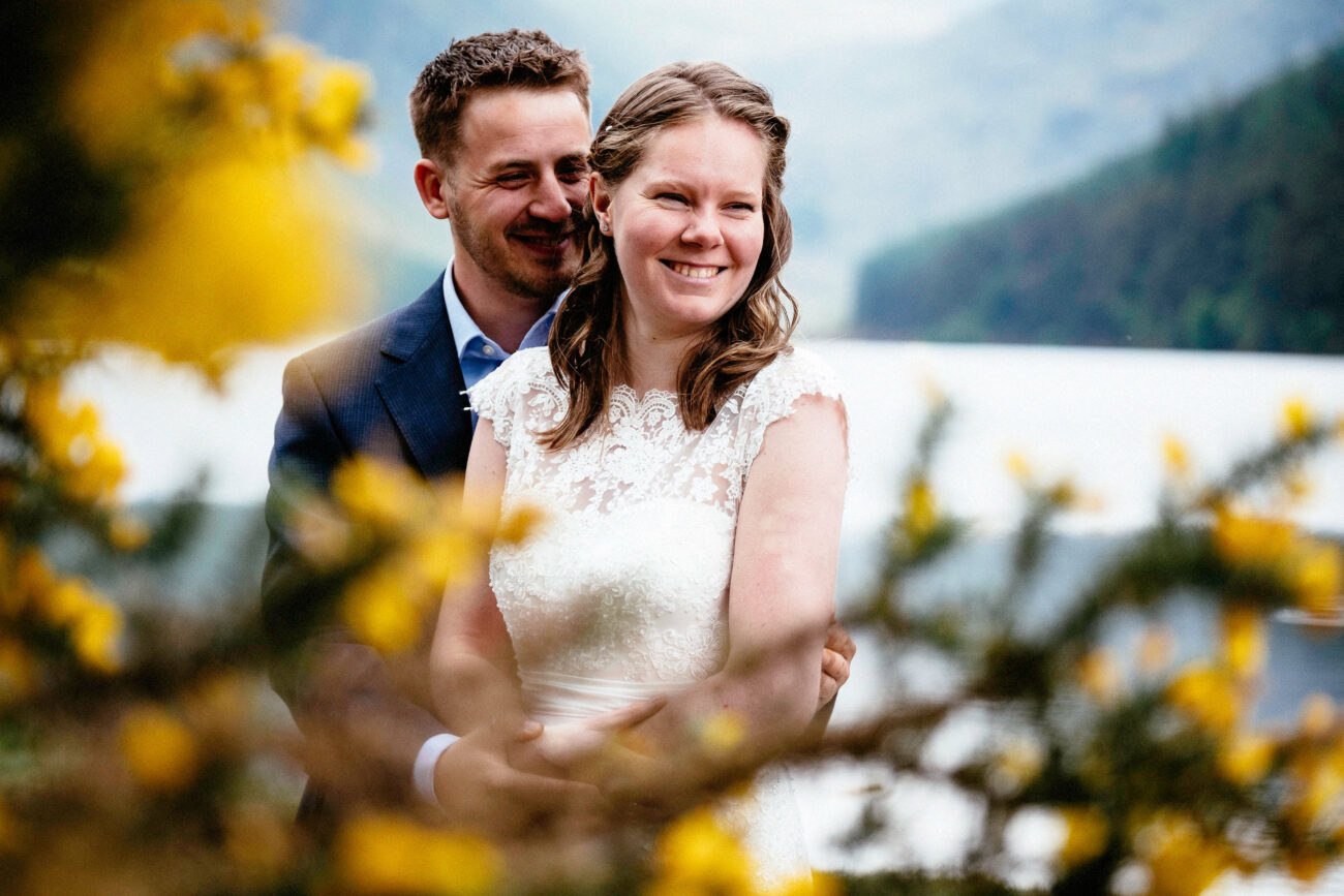 A couple, dressed in formal attire, smiles and embraces near a lake with foliage in the foreground.