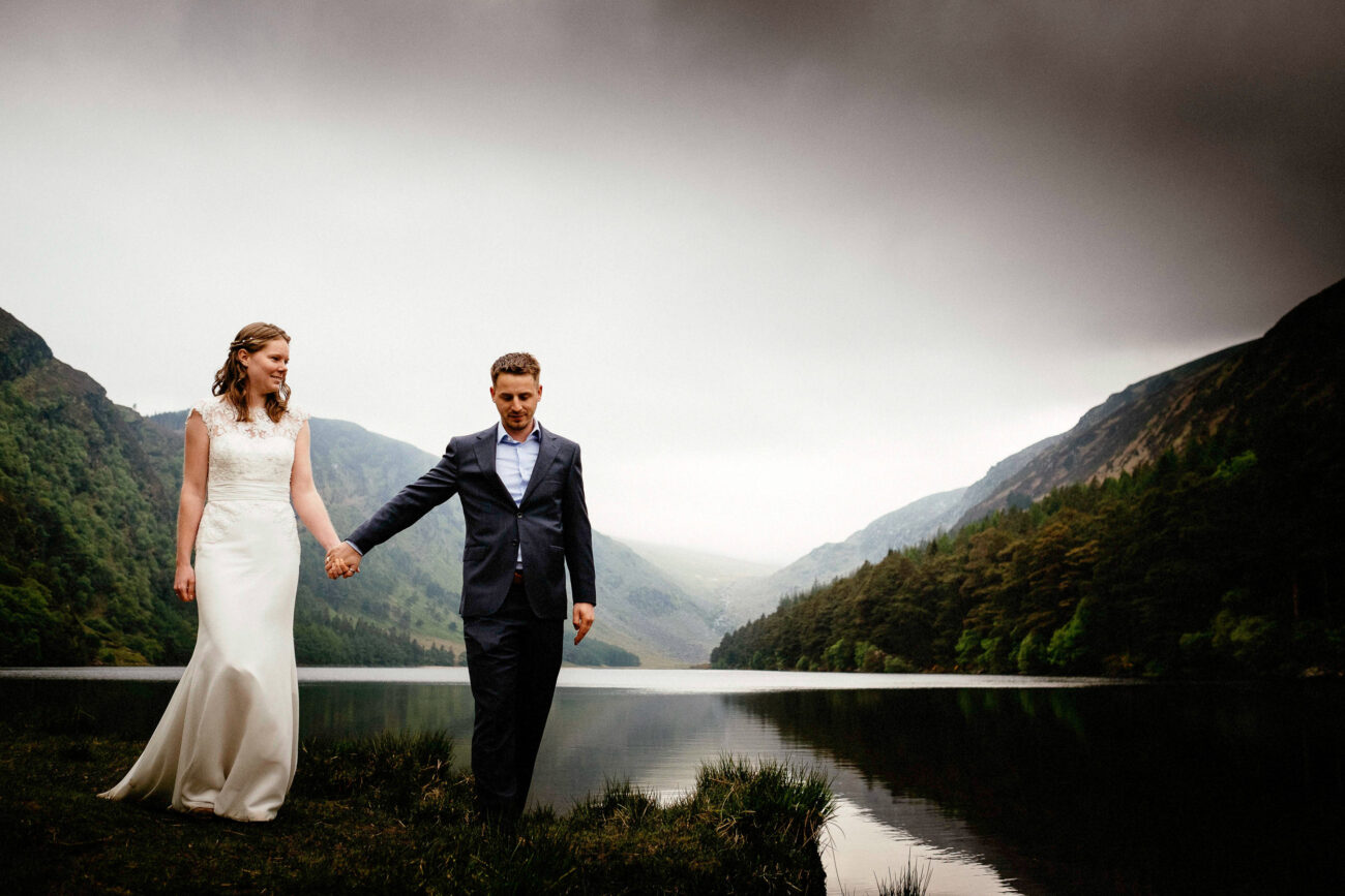 A couple in formal attire holds hands while walking along a lakeside with mountains and a cloudy sky in the background.