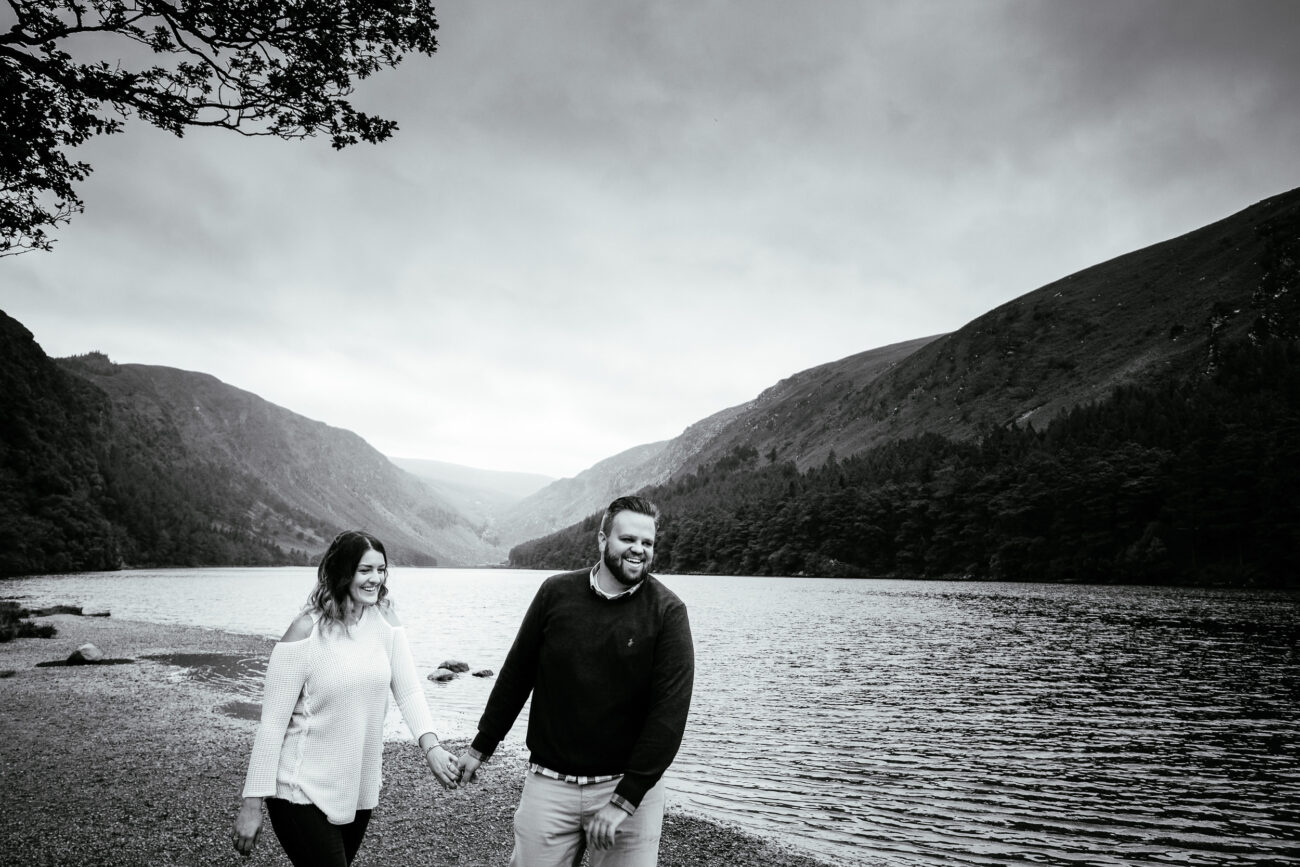 A couple holding hands and walking along the shore of a lake surrounded by hills under a cloudy sky.